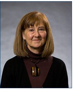 A head-shot of a middle-aged woman with blue eyes and lighter brown hair, she is professionally dressed in a dark maroon turtleneck.